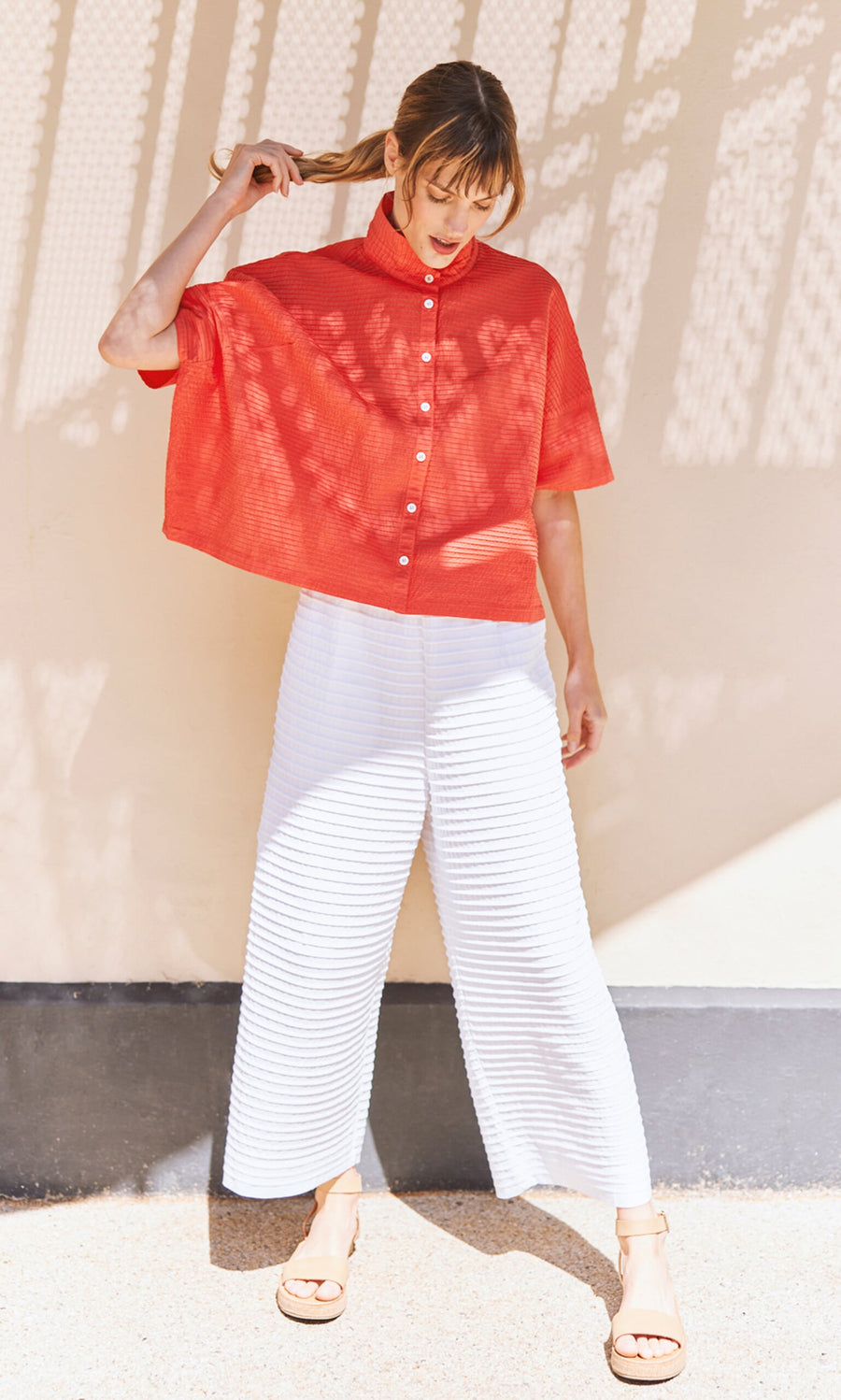 Plümo- Woman in a red top and White Pintucked Pants stands outdoors, holding her ponytail.