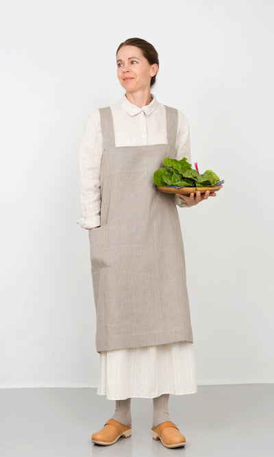 Plümo  Woman in a Linen Pinafore Apron holds a wooden plate of lettuce, standing against a plain white background.