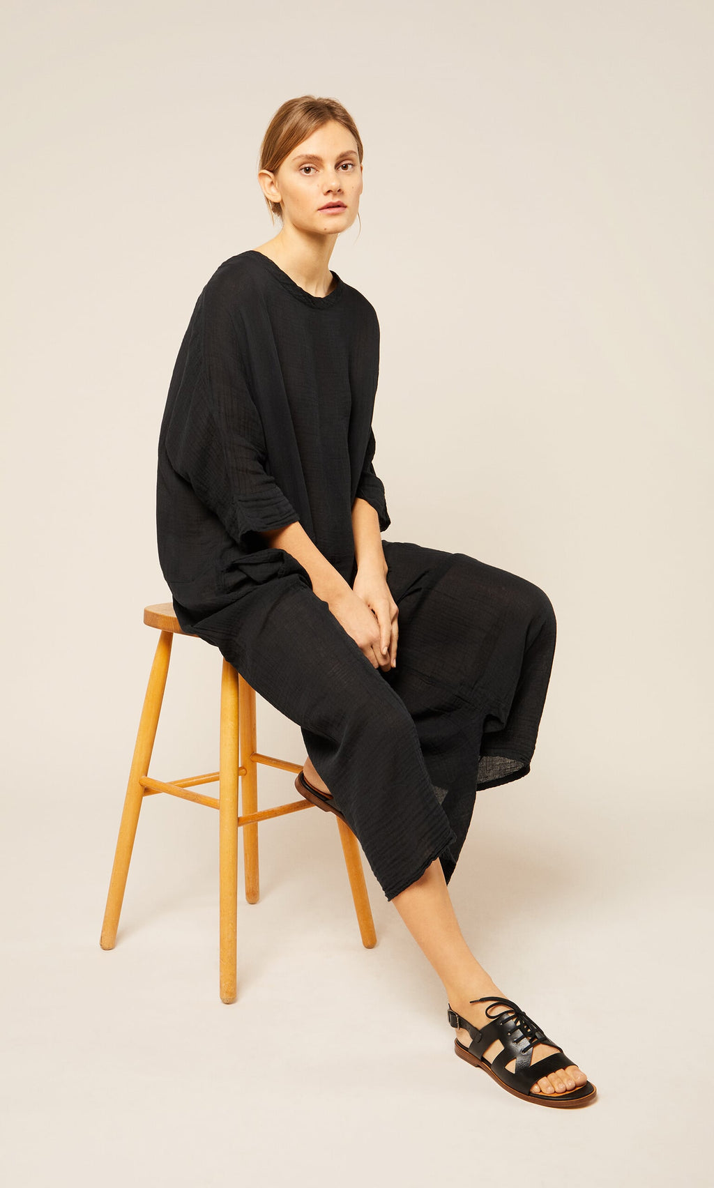 Plümo- Woman in a loose black Culotte Dress sits on a wooden stool against a plain light background, looking at the camera.
