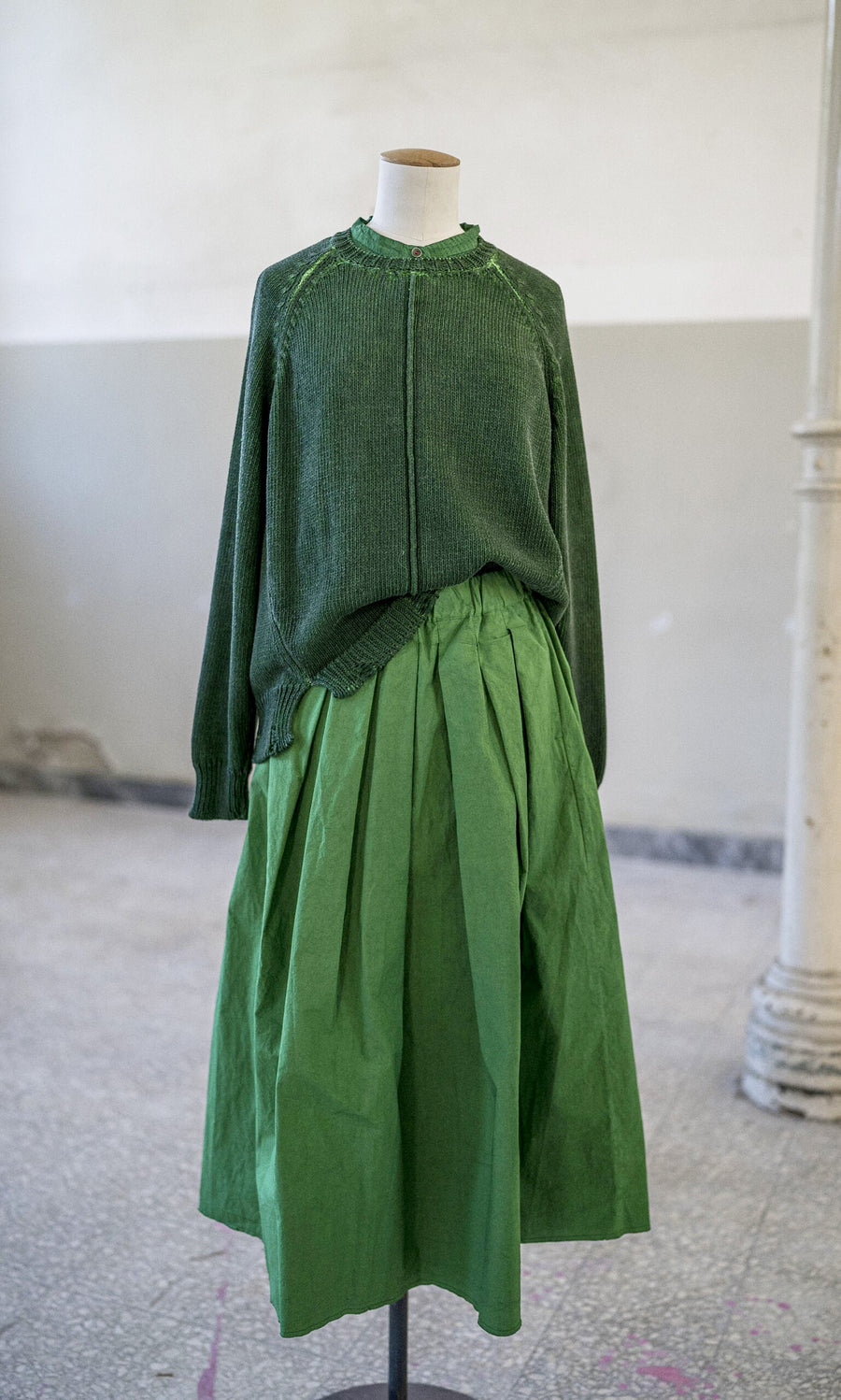 Plümo- Mannequin in a loose dark green sweater and bright green Cadiz Skirt, hand-dyed linen, in a light room.