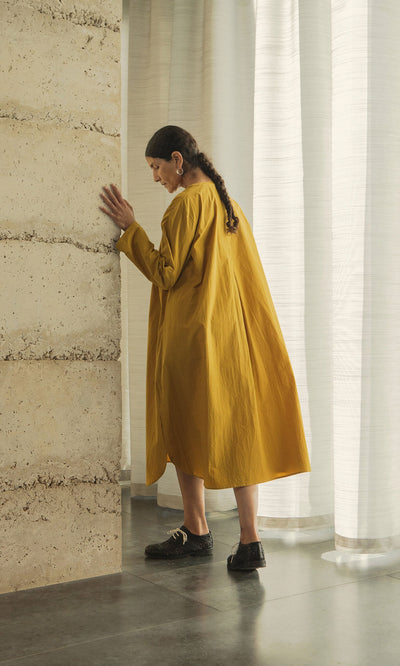Plümo- Someone in a Kaftan Dress stands by a textured wall in a softly lit room with sheer white curtains.