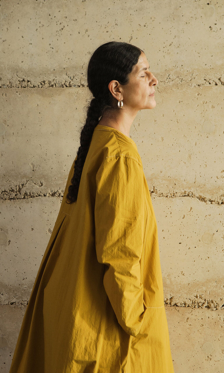 Plümo- Woman in a Kaftan Dress made in London stands in profile, eyes closed, hands in pockets by a beige textured wall.
