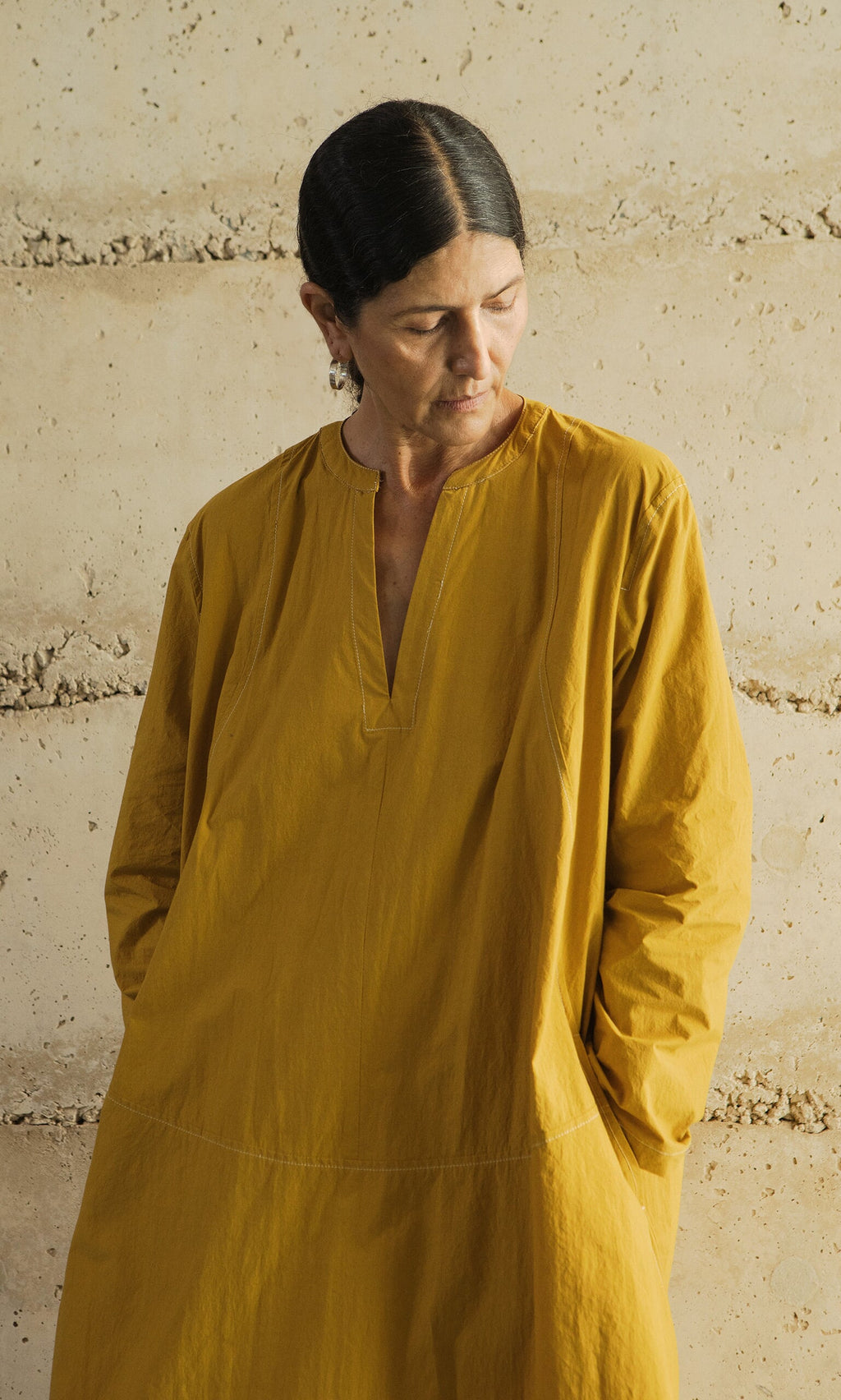 Plümo- A person in a Kaftan Dress stands against a beige wall, looking down with hands in pockets.