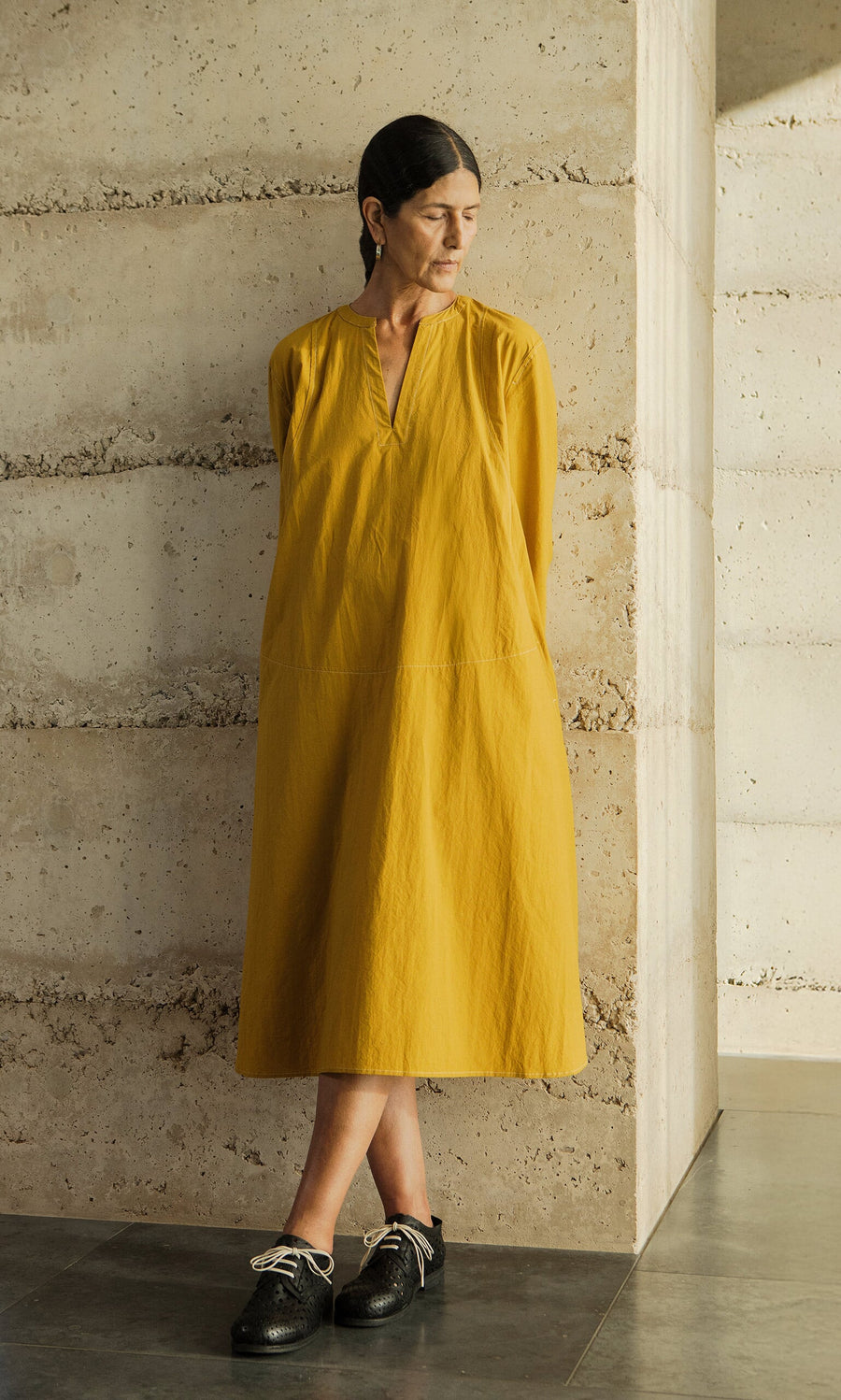 Plümo- Woman in a Kaftan Dress made in London stands by a textured concrete wall, looking to her left.