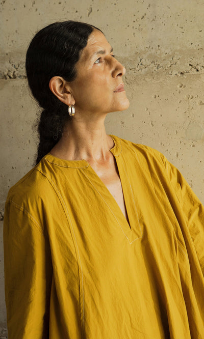 Plümo- A woman in a Kaftan Dress gazes right, standing before a textured beige wall.