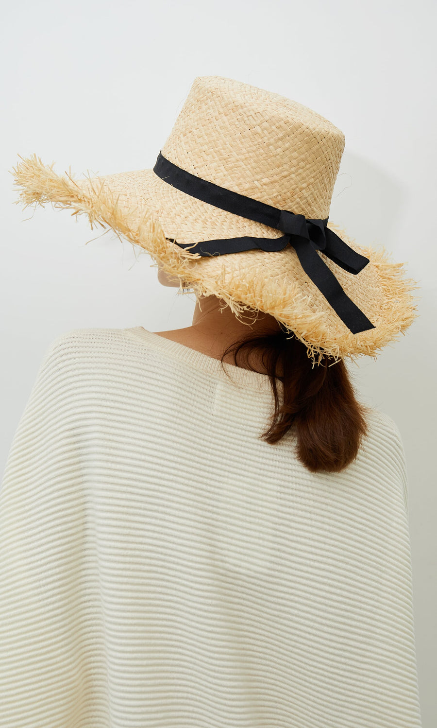 Plümo- Woman in a Frayed Straw Hat with grosgrain ribbon, wearing a cream striped top, facing away.