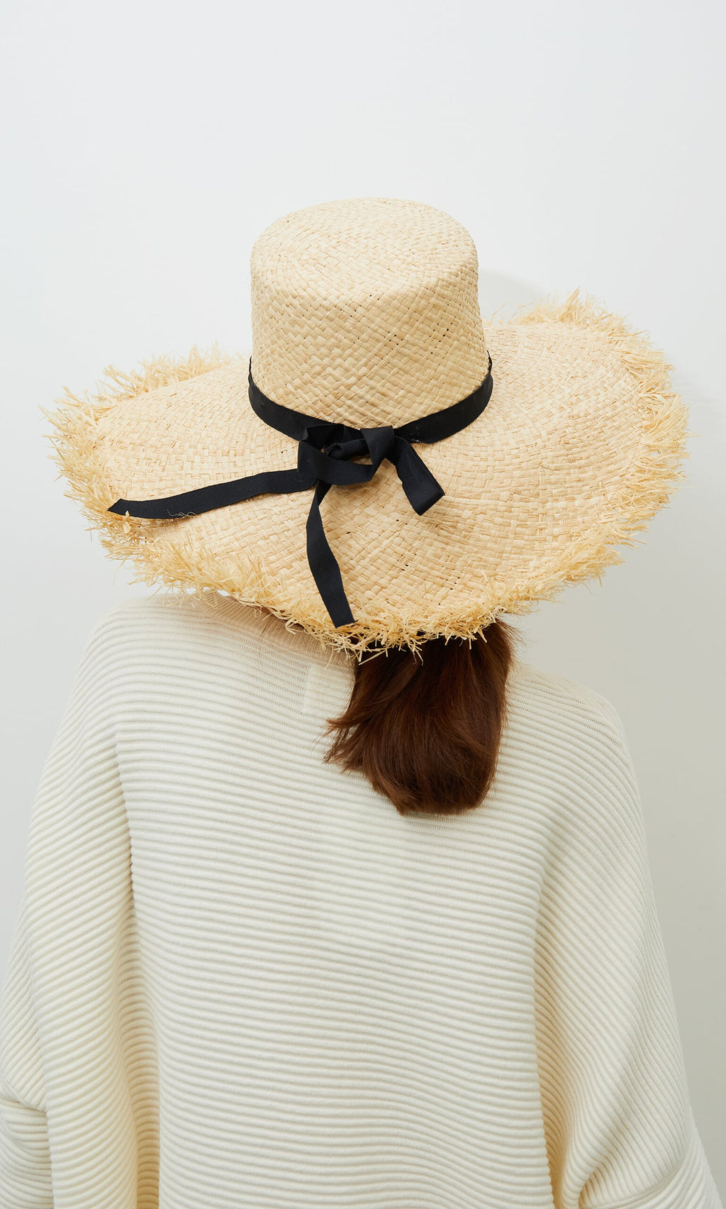 Plümo- Person wearing the Frayed Straw Hat, seen from behind against a plain background.