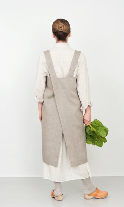Plümo  Woman in a Linen Pinafore Apron stands with back turned, holding leafy greens against a plain white background.