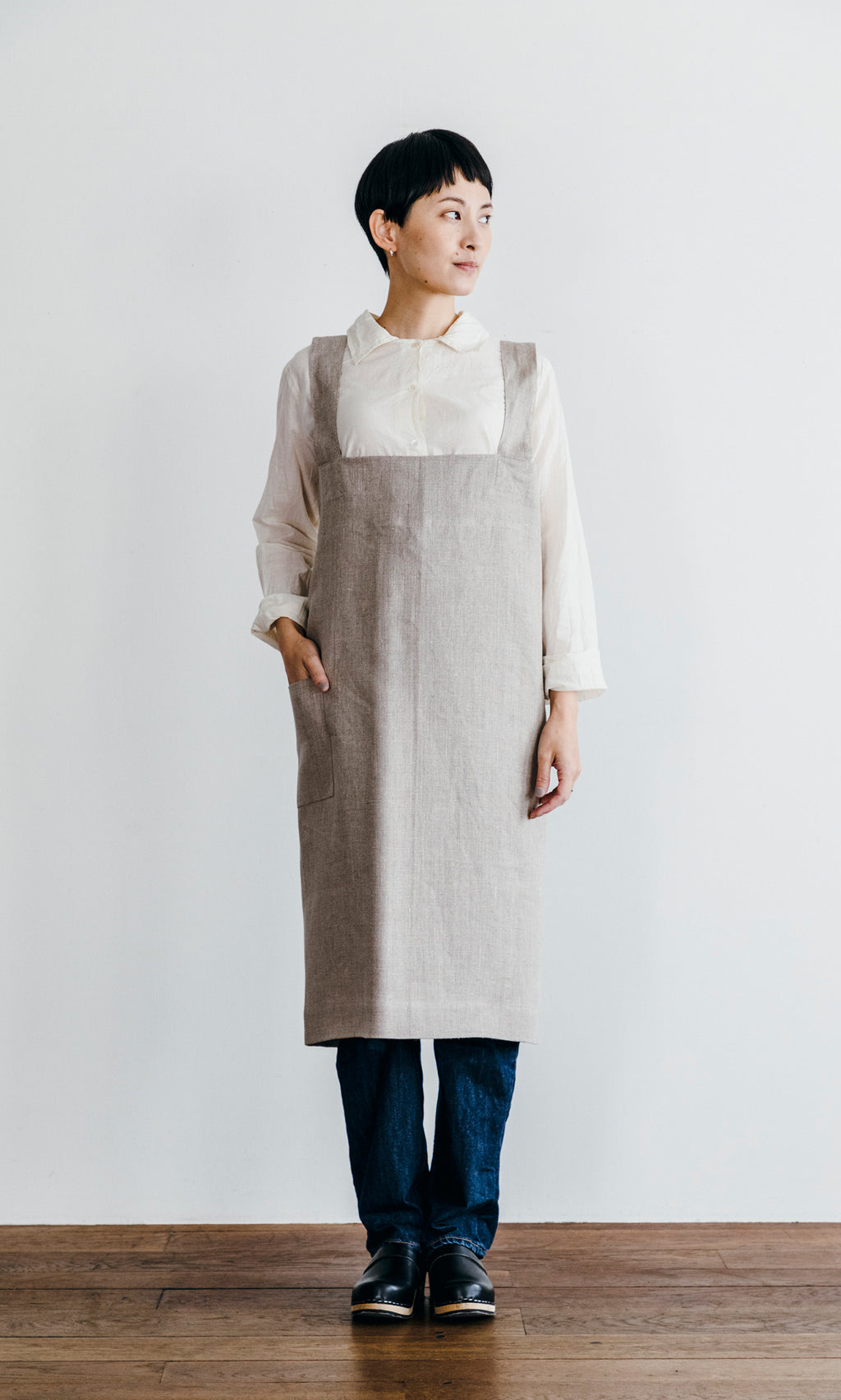 Plümo  Individual in a Linen Pinafore Apron with a deep front pocket stands against a white wall, gazing to the side.