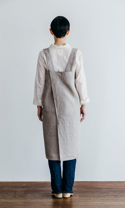 Plümo  Person stands indoors on a wooden floor, wearing a Linen Pinafore Apron over a white shirt and dark pants, facing away.