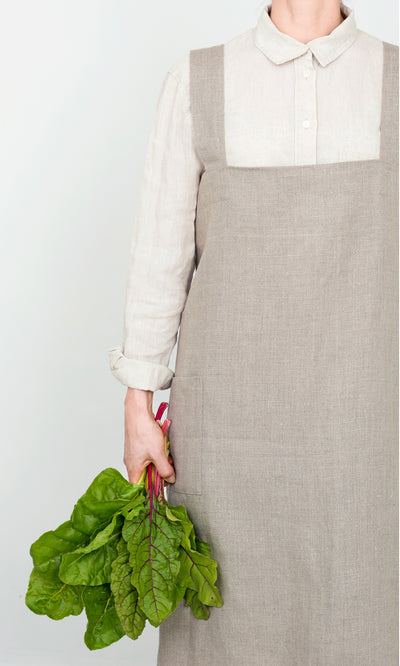 Plümo  Person in a Linen Pinafore Apron holds leafy greens against a simple, light background.