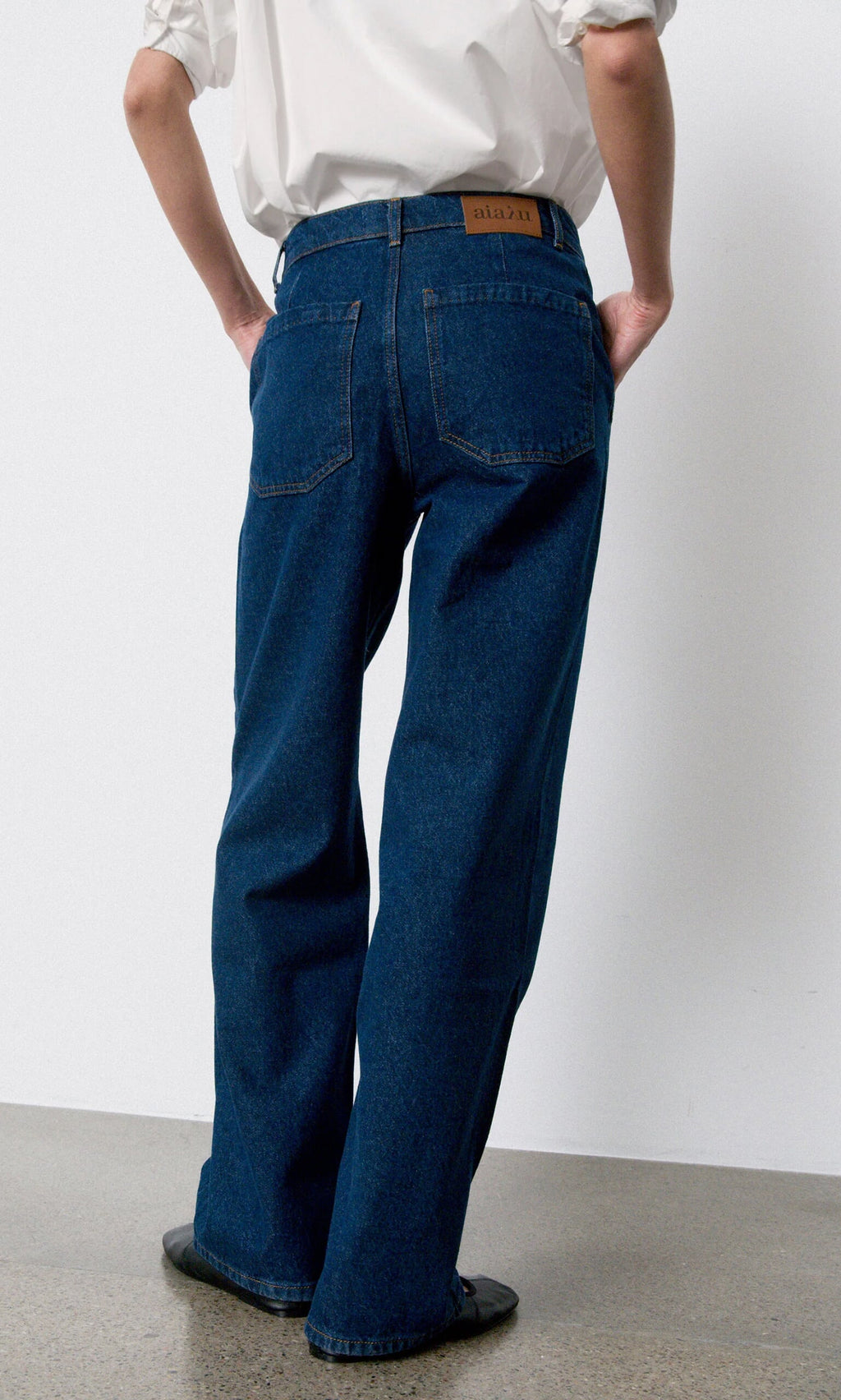 Plümo- Person in a white shirt and Carlo Jeans stands with their back to the camera on a polished floor.