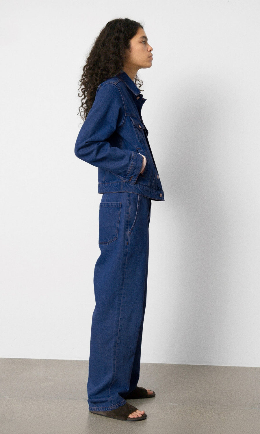 Plümo- Person standing sideways wearing a denim jacket and Carlo Jeans with brown sandals against a plain background.