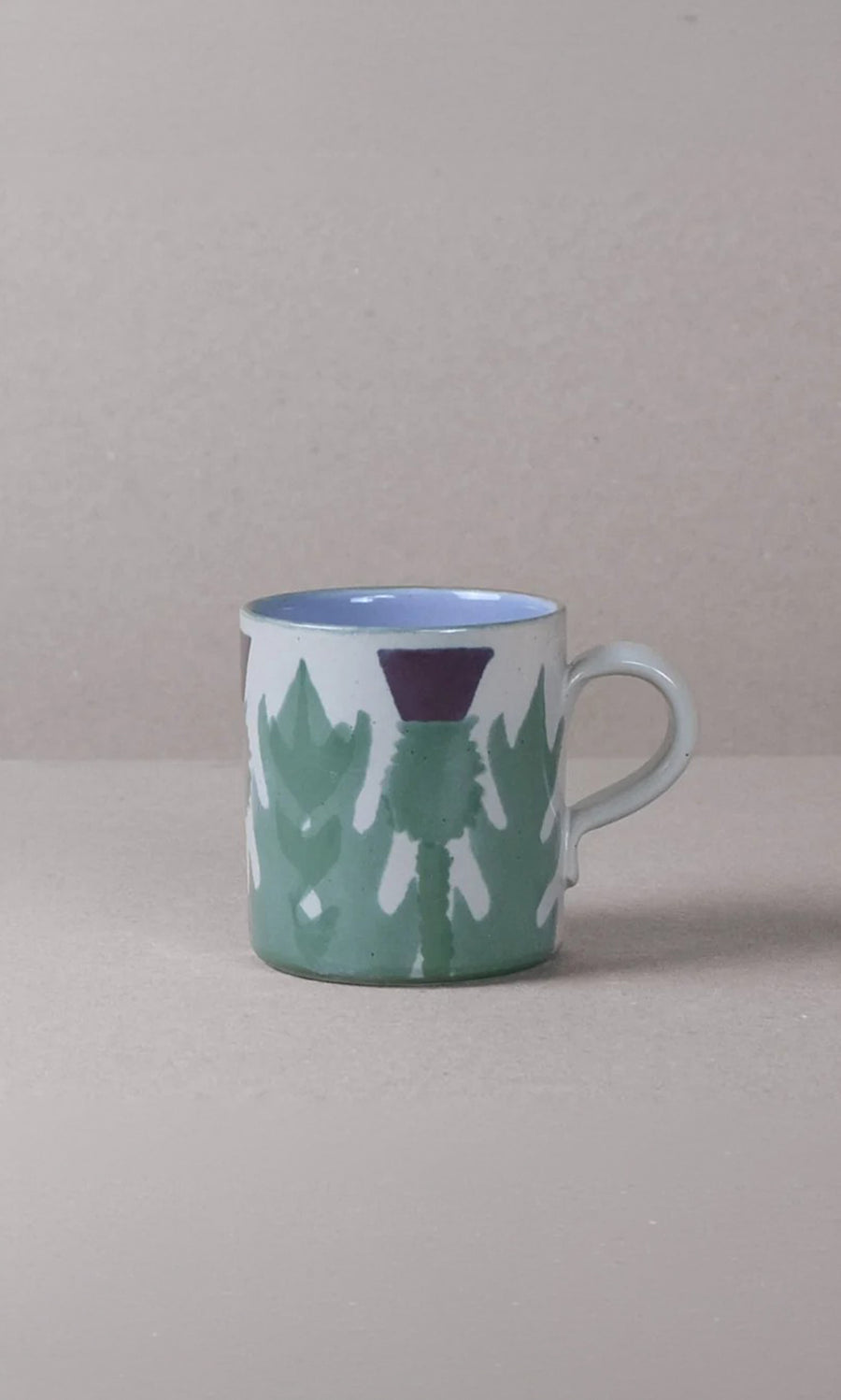 Plümo  The Thistle Mug, hand-painted with a green leaf and purple flower, rests on a beige surface with a beige background.