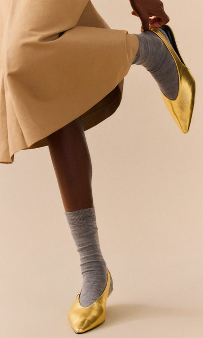 Plümo  A person in a tan skirt wears slate grey Alpaca Socks and shiny gold ballet flats, one foot lifted.