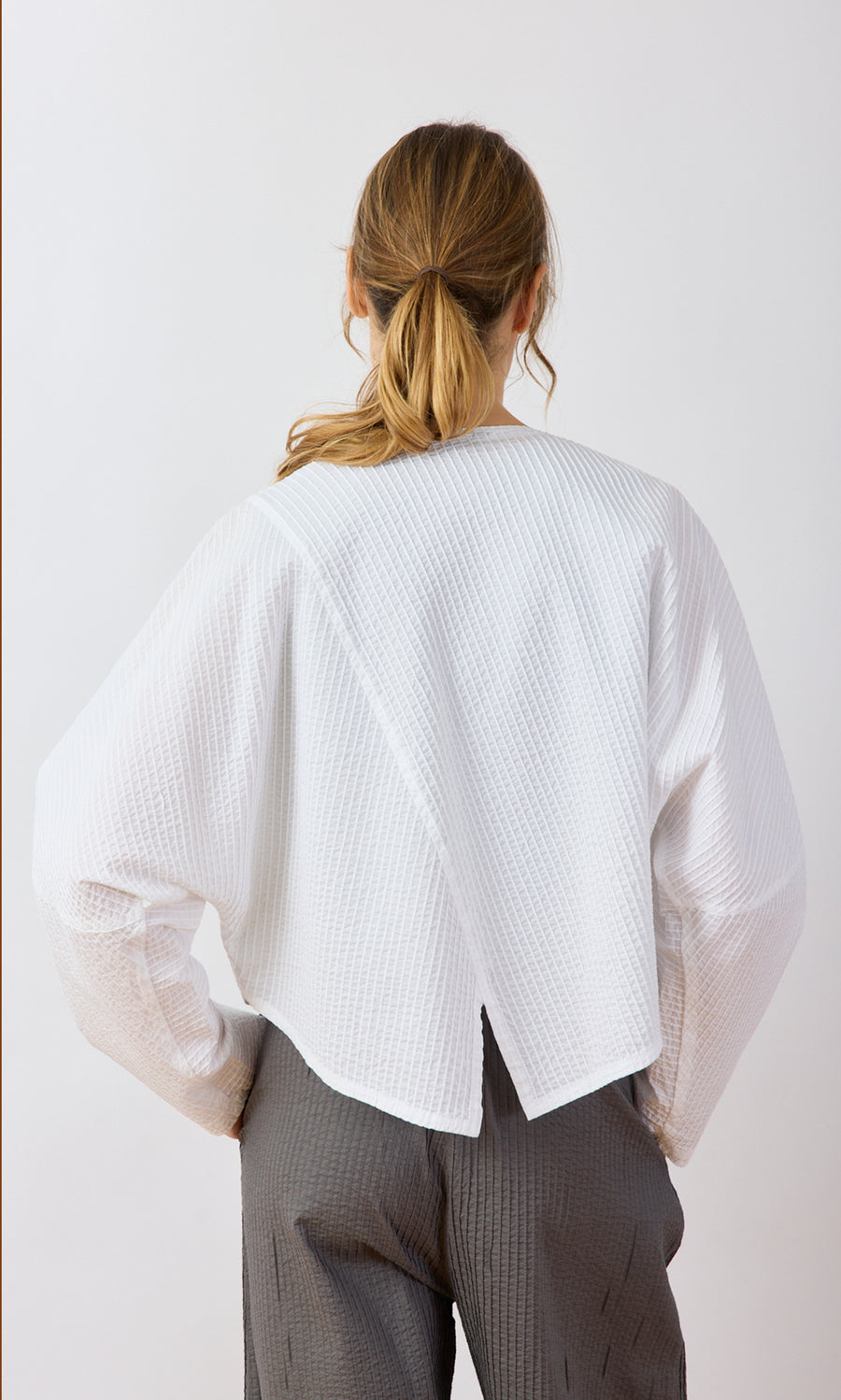 Plümo- Woman facing away in an Orna Pleat Jacket and gray pants, hair styled in a loose ponytail.
