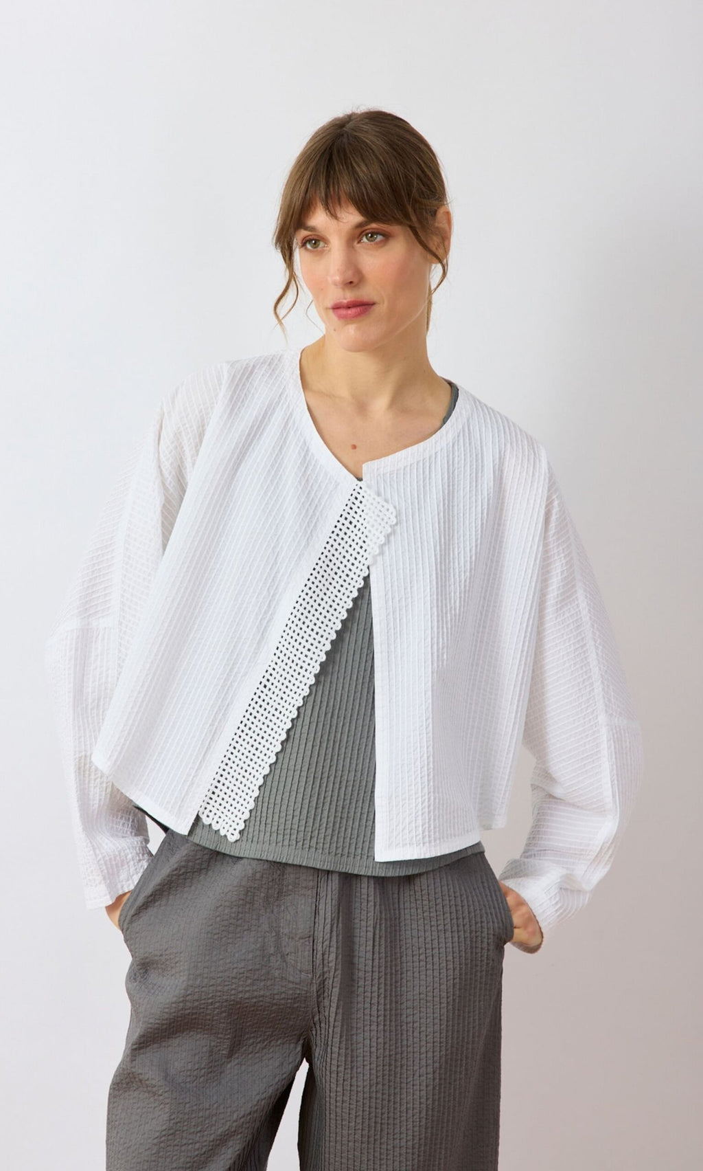 Plümo- Woman wearing the Orna Pleat Jacket over a gray top and pants, standing against a light background.