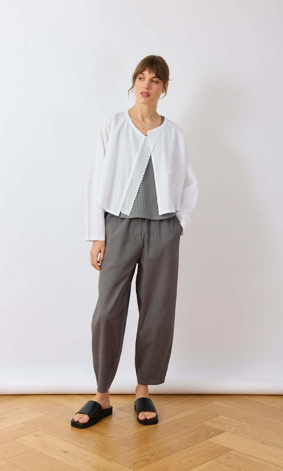 Plümo- Woman on wooden floor wears Orna Pleat Jacket, gray pants, and black slides.
