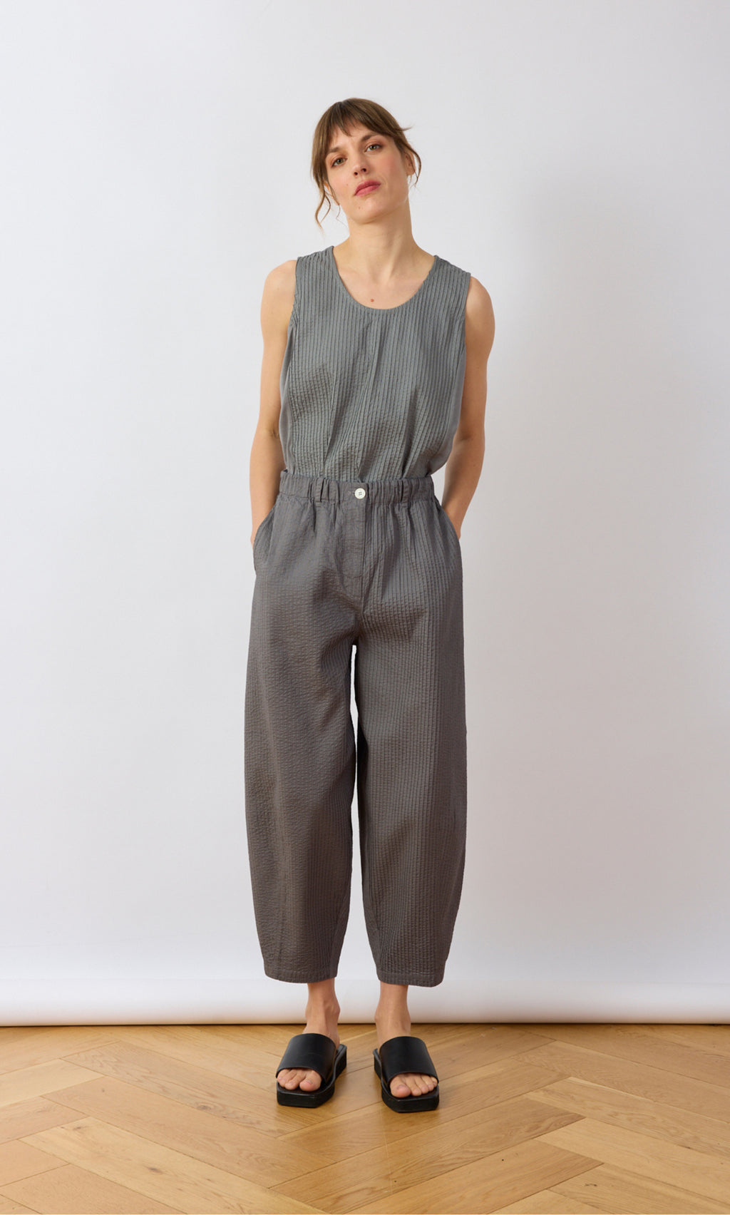 Plümo- Woman in a sleeveless gray top and Haru Grey Pants stands with hands in pockets against a white background.