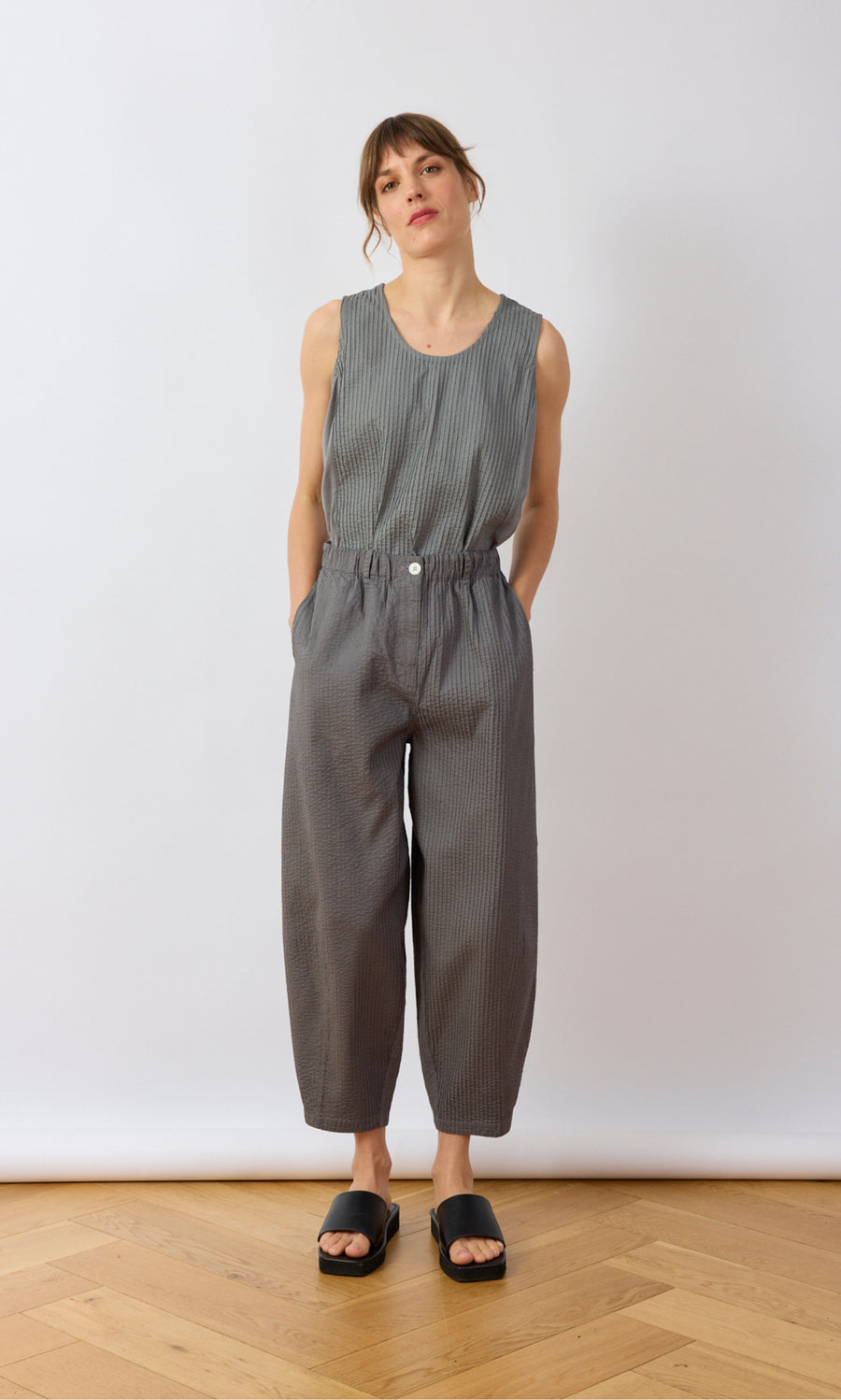 Plümo- Woman in a sleeveless gray top and Haru Grey Pants stands with hands in pockets against a white background.