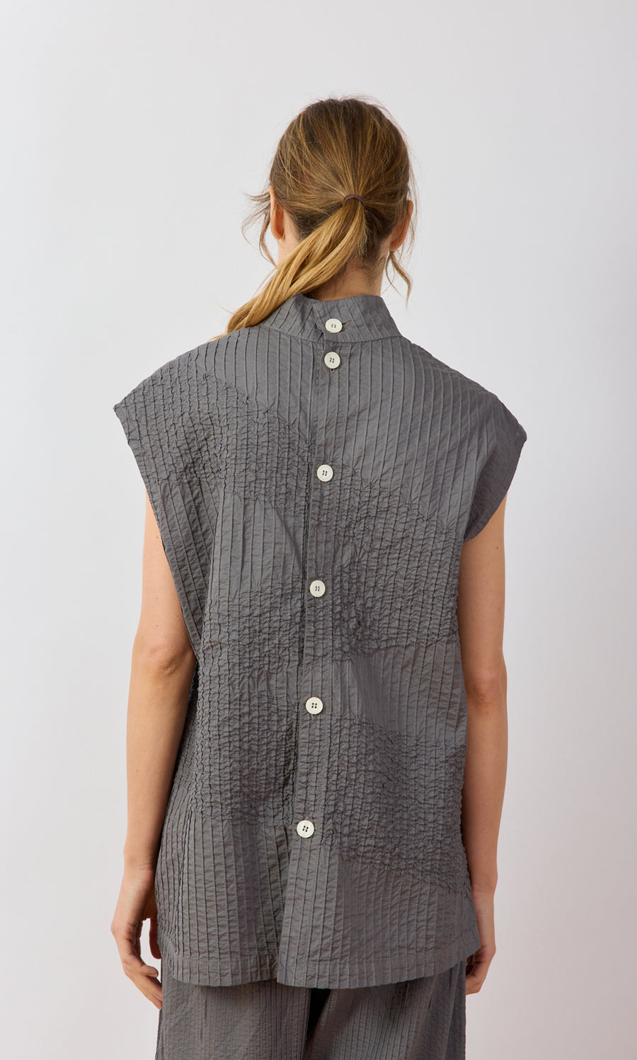 Plümo- Woman with hair in a low ponytail wears the Minato Top, a gray pintucked Yacco Maricard design with large white buttons.