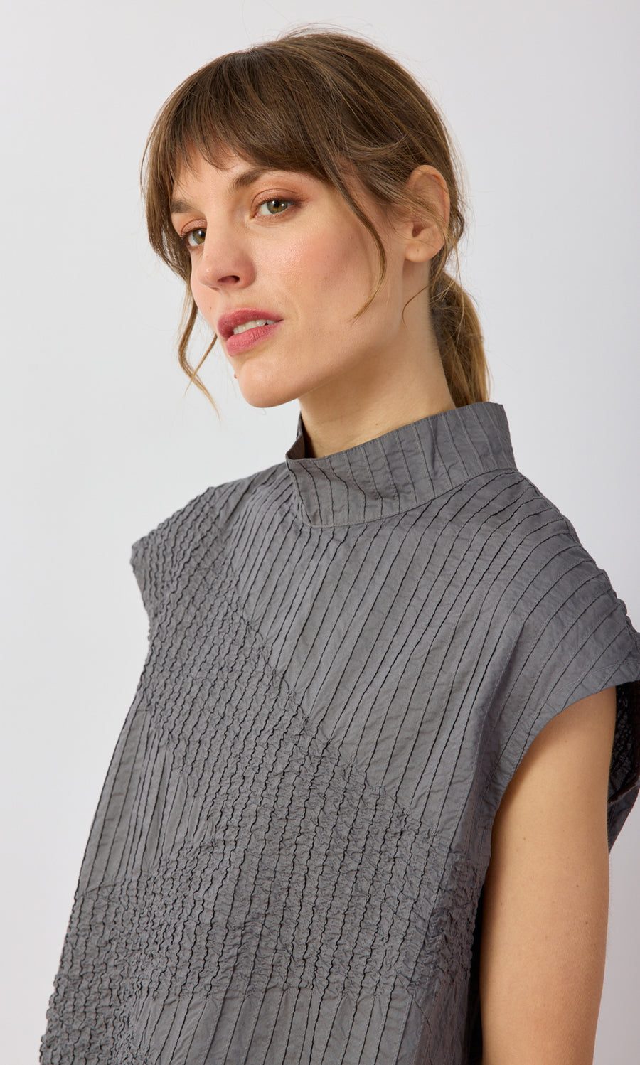 Plümo- Woman with light brown hair in a ponytail, wearing a gray Minato Top, looking slightly to the side.