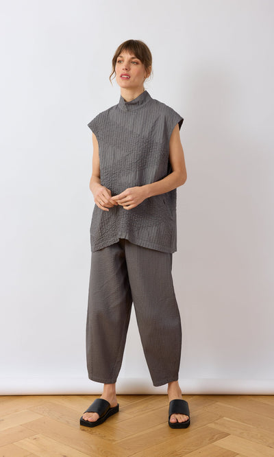 Plümo- Woman wearing a grey textured Minato Top and pants with black slides, standing on a wooden floor against a white wall.