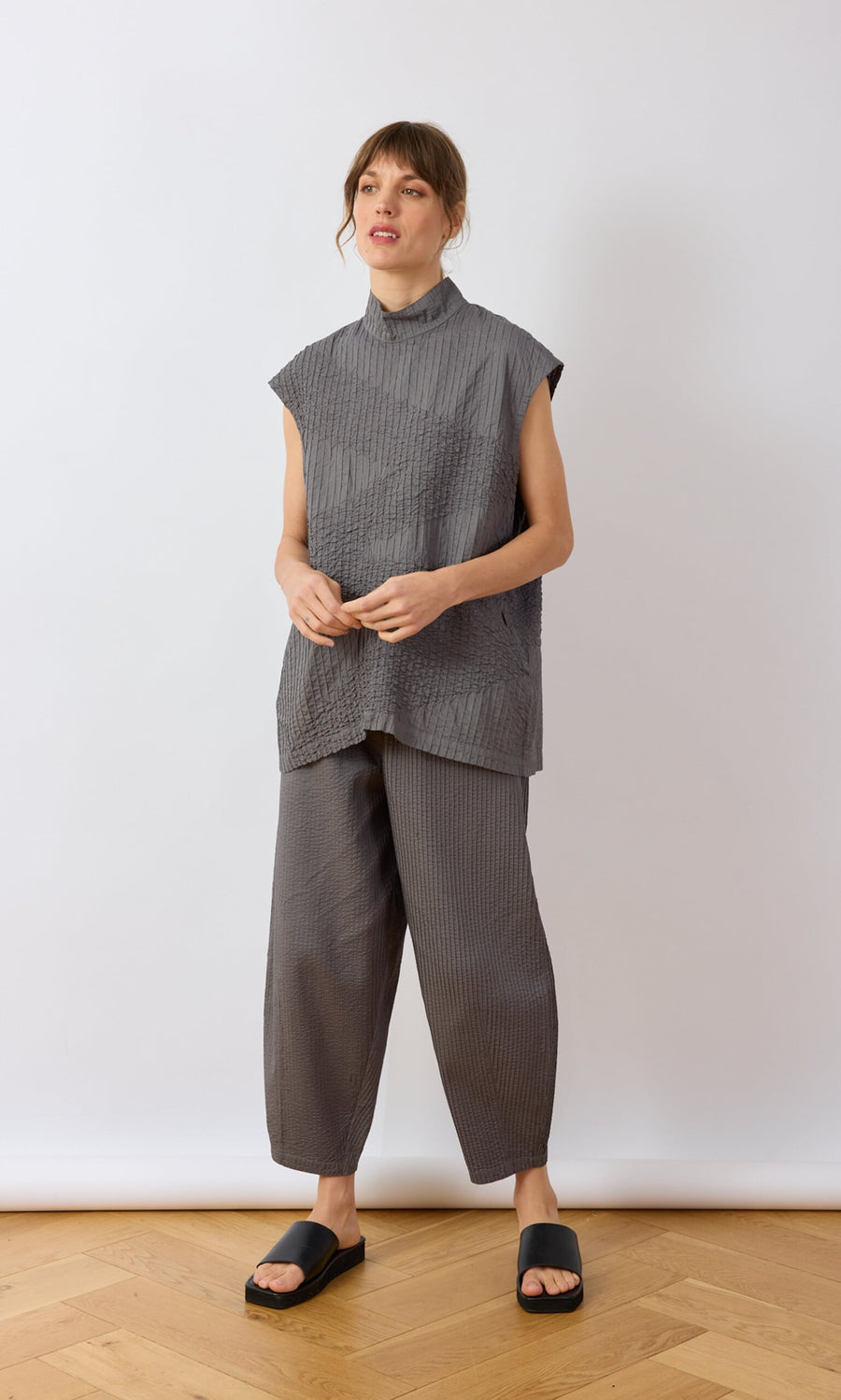 Plümo- Woman wearing a grey textured Minato Top and pants with black slides, standing on a wooden floor against a white wall.
