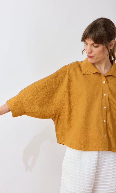 Plümo- Woman in the Karashi Shirt—loose mustard yellow with wide sleeves—paired with white textured pants.