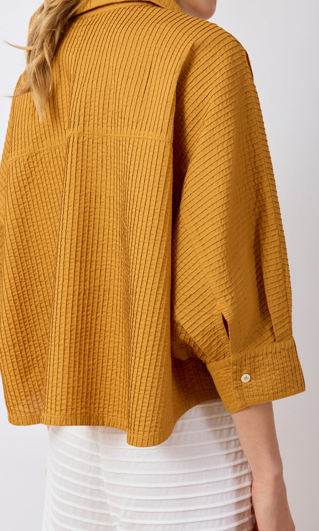 Plümo- A person in a textured mustard yellow Karashi Shirt and white skirt, shown from the back and side.