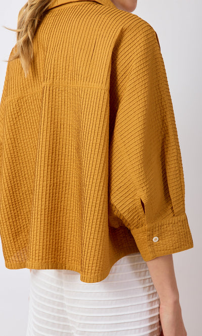 Plümo- A person in a textured mustard yellow Karashi Shirt and white skirt, shown from the back and side.