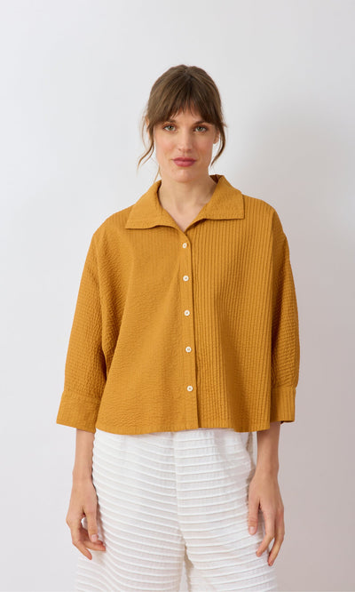 Plümo- Woman wearing a mustard yellow Karashi Shirt with white pleated pants, standing against a plain background.