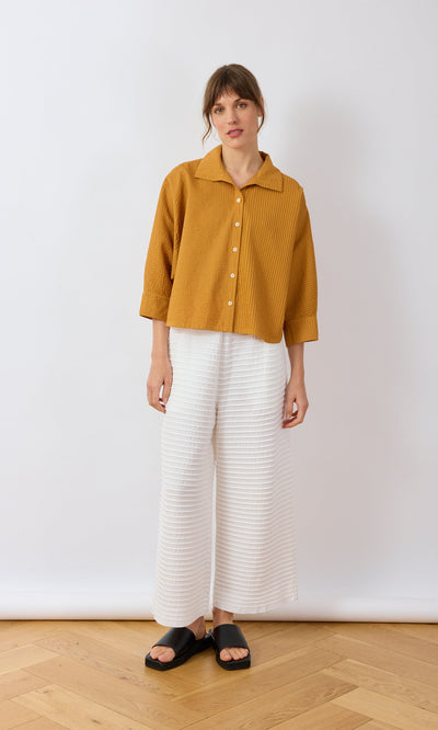 Plümo- Woman in a Karashi Shirt, white wide-leg pants, and black sandals against a white background.