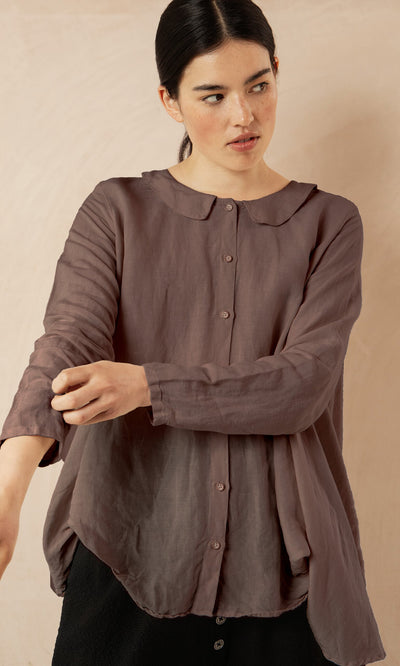 Plümo- Woman in the Beeson Shirt - Mushroom, with Peter Pan collar, rolls up her sleeve against a beige background.