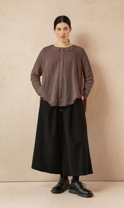 Plümo- Woman stands against a beige wall in the Beeson Shirt - Mushroom, black wide-leg pants, and black shoes, hands in pockets.