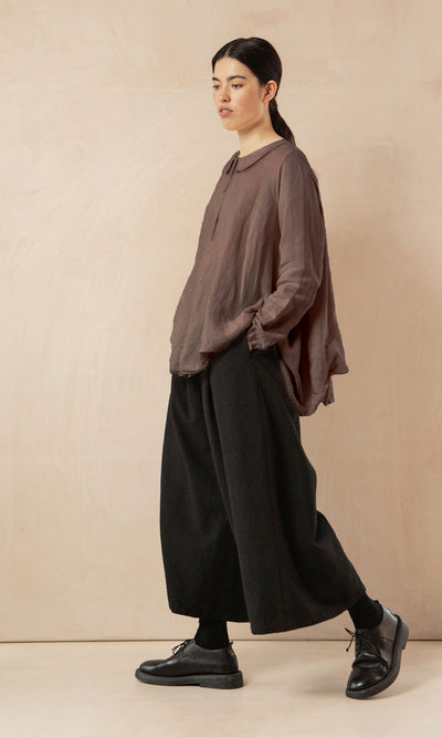 Plümo- Woman in the Beeson Shirt - Mushroom and wide black pants, hands in pockets, walking against a beige background.