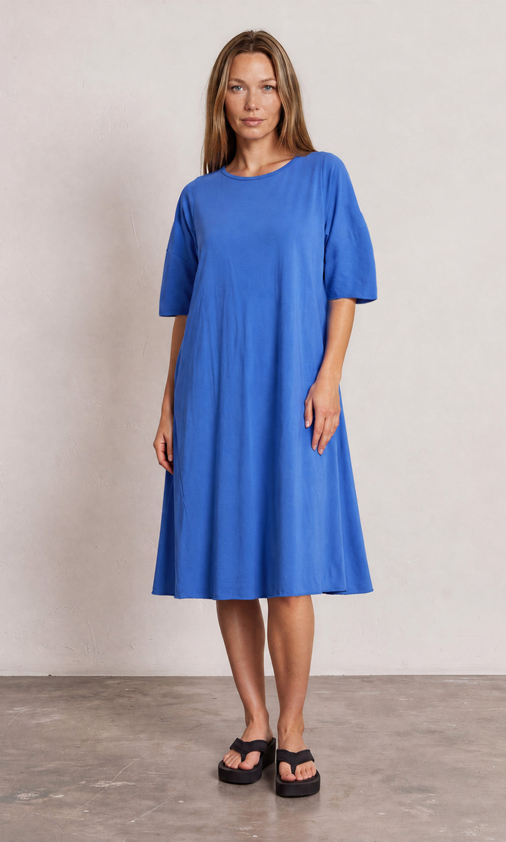 Plümo  Woman in a loose-fitting Bevell Dress and black sandals stands on a gray floor against a plain backdrop.
