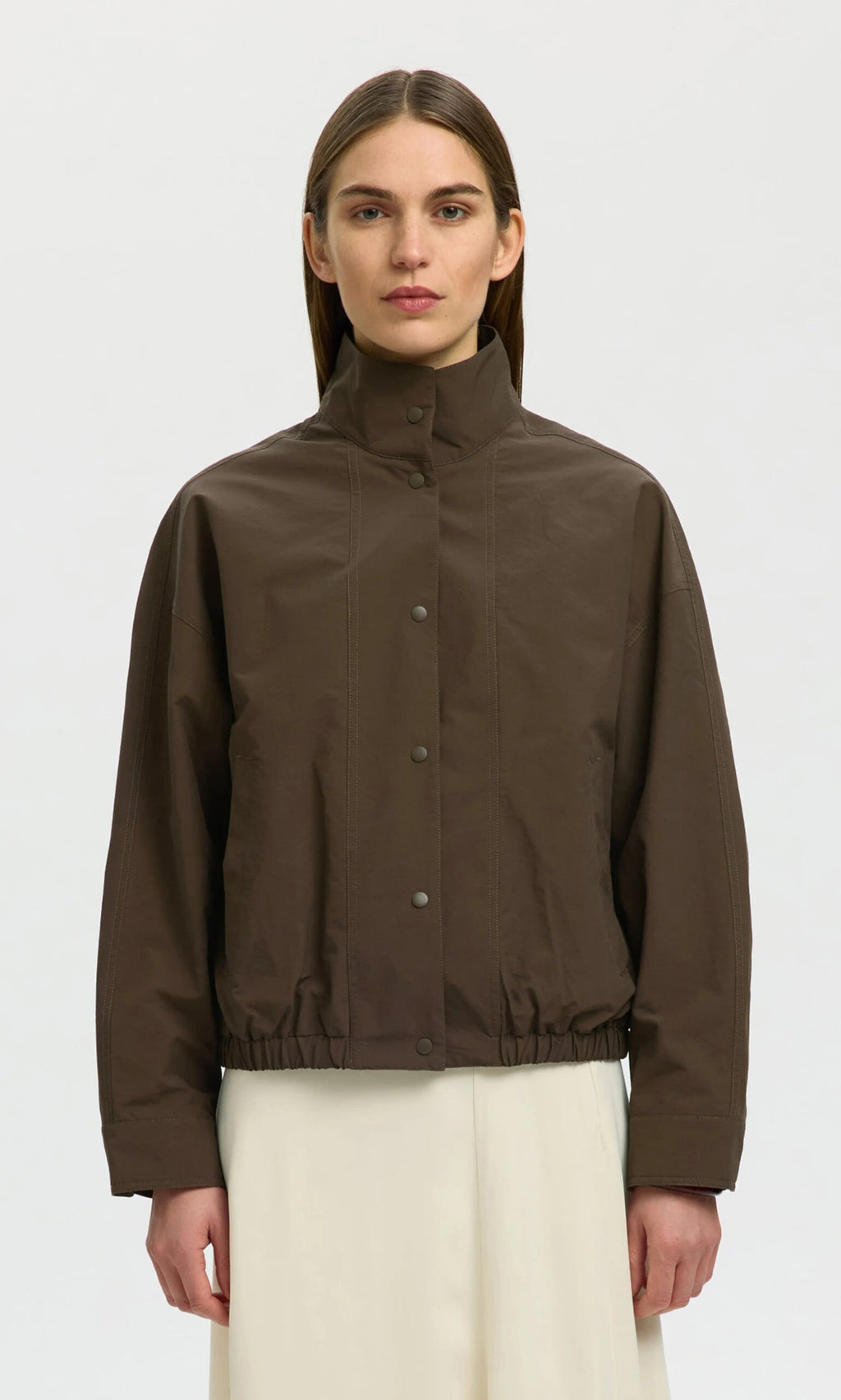 Plümo- A woman models a brown, water-resistant Bomber Jacket with snap buttons and elastic hem, paired with a white skirt.
