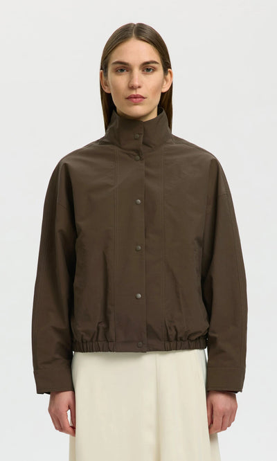 Plümo- A woman models a brown, water-resistant Bomber Jacket with snap buttons and elastic hem, paired with a white skirt.