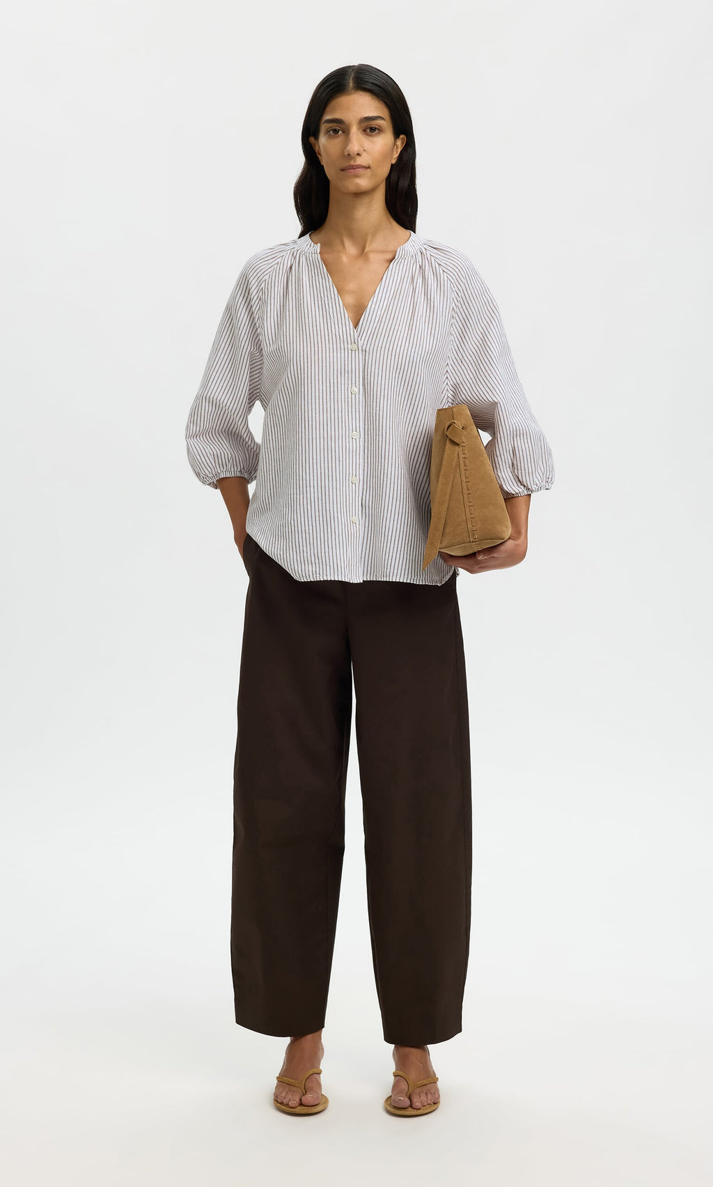 Plümo- Woman in a relaxed fit Brisley Blouse, dark wide-leg pants, sandals, and tan bag standing against a white background.