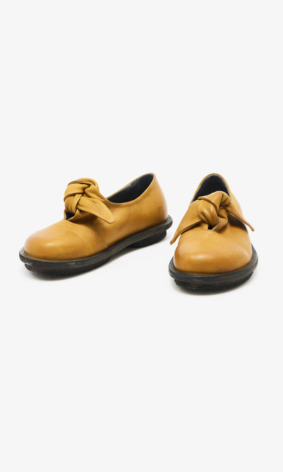 Plümo  Amber Leather Shoes: Mustard yellow flats with black soles, knotted bows, Danish-inspired, on white background.