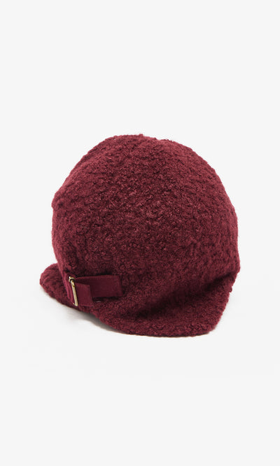Plümo  The Velo Wool Cap is a maroon boucle hat with ear flaps, a short brim, and a small decorative buckle.