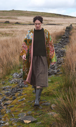 Plümo  Person in an Aurelie Velvet Coat, green sweater, skirt, and boots walks along a rocky path through grassy moorland.