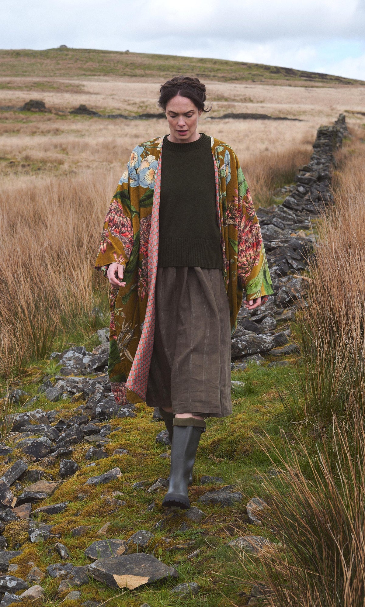 Plümo  Person in an Aurelie Velvet Coat, green sweater, skirt, and boots walks along a rocky path through grassy moorland.