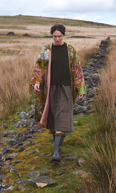 Plümo  Person in an Aurelie Velvet Coat, green sweater, skirt, and boots walks along a rocky path through grassy moorland.