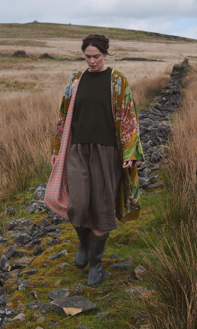 Plümo  A woman in the Aurelie Velvet Coat and boots walks on a rocky path through tall grass in a rural landscape.