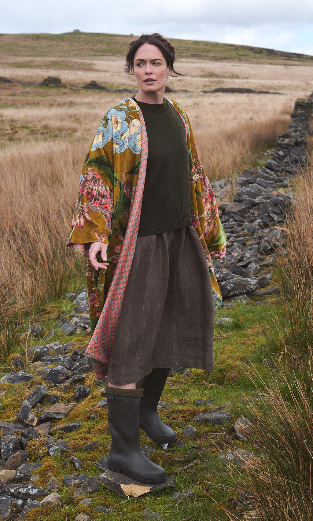 Plümo  Woman in an Aurelie Velvet Coat stands on a grassy path by a stone wall, styled with skirt and boots.