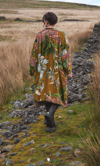 Plümo  Person in an Aurelie Velvet Coat walks on a rocky path through grassy fields bordered by stone walls.