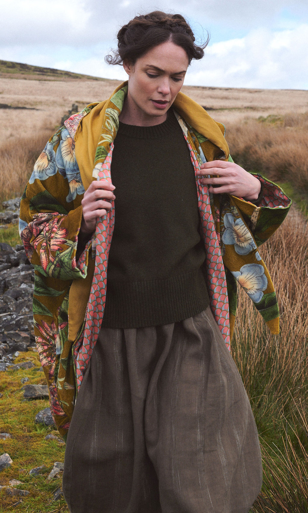 Plümo  A woman in the Aurelie Velvet Coat walks outdoors in a grassy, rocky landscape under a cloudy sky.
