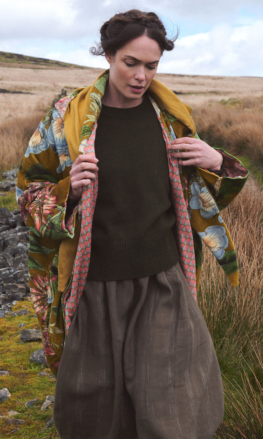 Plümo  A woman in the Aurelie Velvet Coat walks outdoors in a grassy, rocky landscape under a cloudy sky.